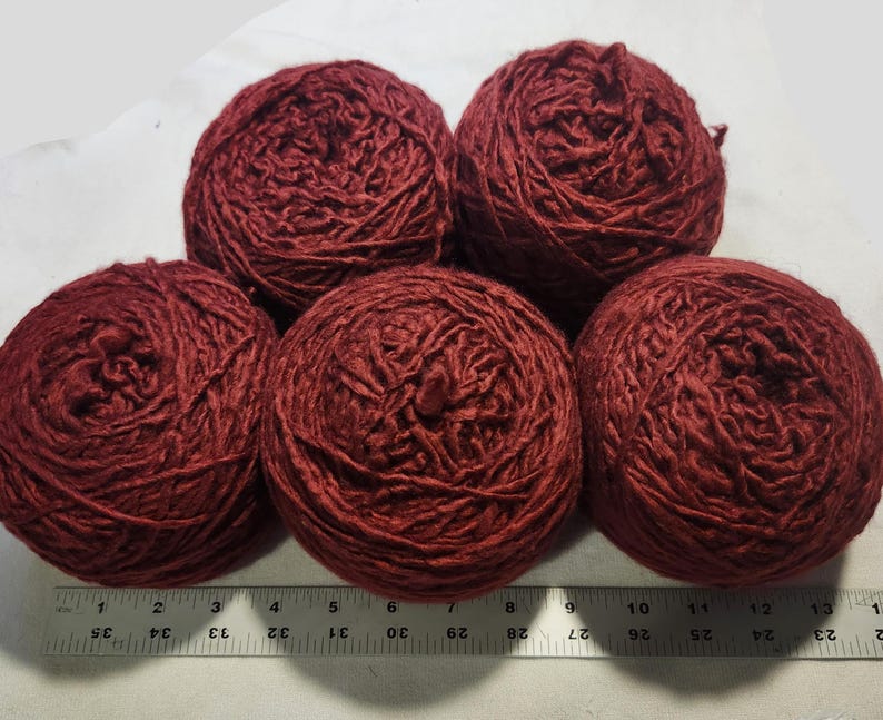 May include: Five round balls of deep red yarn arranged in a cluster. A ruler is positioned along the bottom edge, showing the width of the yarn balls spans approximately 13 inches across. The yarn has a textured, slightly twisted appearance.