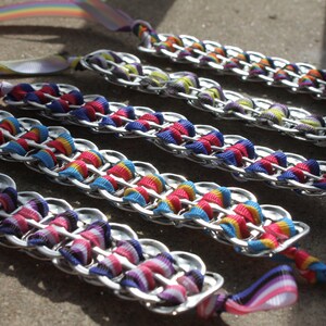 May include: Several handmade bracelets made from silver-colored metal tabs and colorful ribbons. The ribbons are woven through the tabs in a chain-like pattern, with rainbow-colored ribbons and solid colors. The bracelets are laid out on a gray surface.