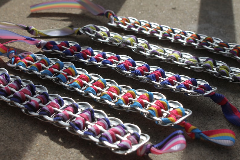 May include: Several handmade bracelets made from silver-colored metal tabs and colorful ribbons. The ribbons are woven through the tabs in a chain-like pattern, with rainbow-colored ribbons and solid colors. The bracelets are laid out on a gray surface.