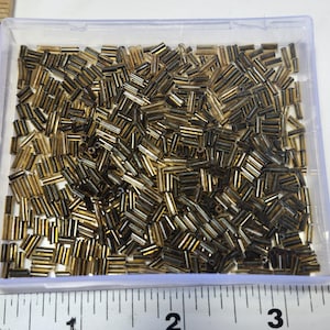 May include: A clear plastic container filled with numerous small, cylindrical beads. The beads are primarily gold and bronze in color, with a slightly translucent appearance. The container is placed next to a ruler, indicating the beads' small size, suitable for jewelry making.