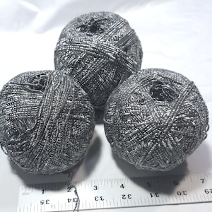May include: Three spherical balls of metallic silver yarn arranged in a triangular formation. A white ruler is positioned at the bottom, showing the balls measure approximately 2.5 inches in diameter each, with a textured, reflective surface.