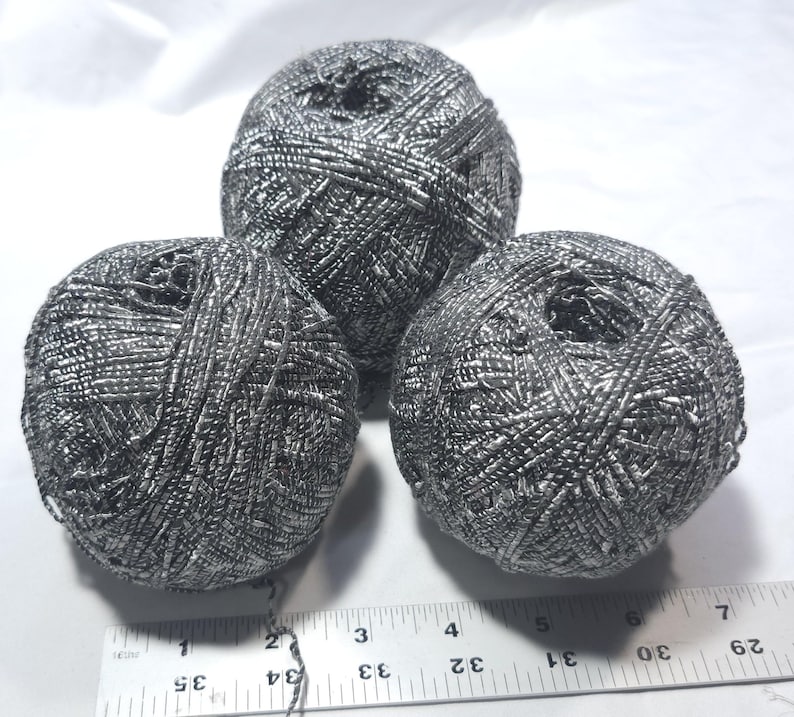 May include: Three spherical balls of metallic silver yarn arranged in a triangular formation. A white ruler is positioned at the bottom, showing the balls measure approximately 2.5 inches in diameter each, with a textured, reflective surface.