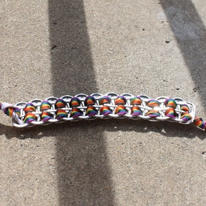 May include: A handmade bracelet featuring a chainmail design made from silver-colored metal. The bracelet is accented with rainbow-colored ribbon woven through the links. The ribbon is also used as the bracelet's ties.