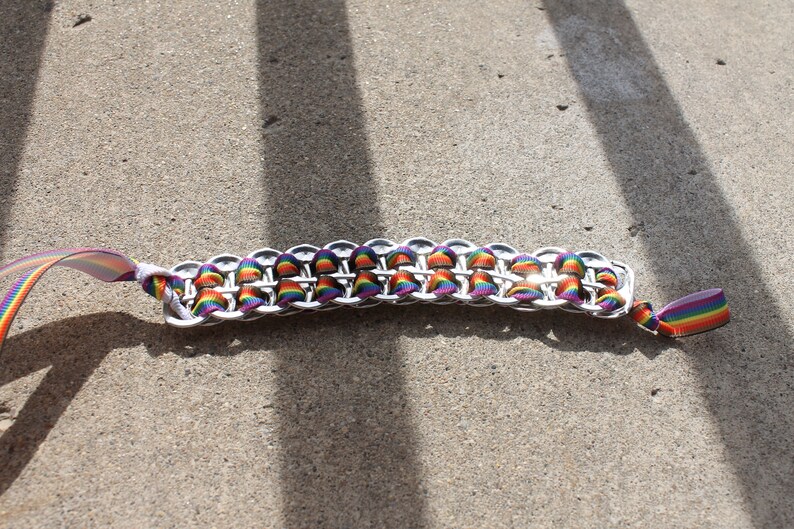May include: A handmade bracelet featuring a chainmail design made from silver-colored metal. The bracelet is accented with rainbow-colored ribbon woven through the links. The ribbon is also used as the bracelet's ties.
