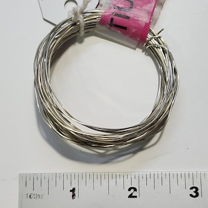May include: Coiled silver-colored wire, likely for jewelry making or crafts. A pink tag is attached to the wire. A ruler with inch measurements is in the foreground.