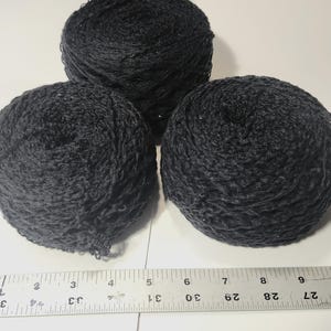 May include: Three round balls of black yarn with a textured, net-like appearance. A metal ruler is positioned at the bottom, showing the yarn balls span approximately 9 inches in width across the bottom edge of the arrangement.