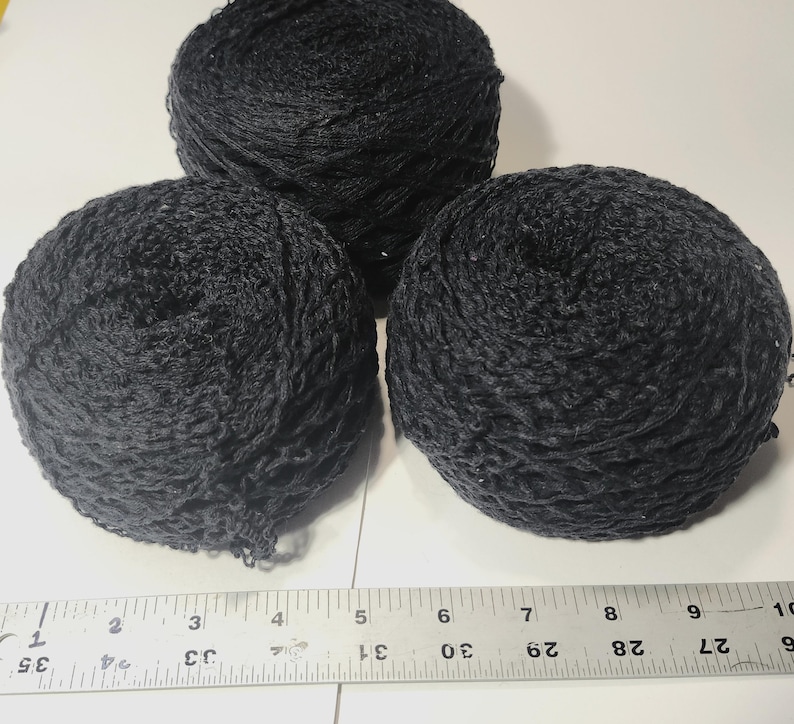 May include: Three round balls of black yarn with a textured, net-like appearance. A metal ruler is positioned at the bottom, showing the yarn balls span approximately 9 inches in width across the bottom edge of the arrangement.
