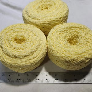 May include: Three round cakes of pale yellow yarn arranged in a triangular formation. A metal ruler is positioned at the base, showing that each yarn cake has a diameter of approximately 3.5 inches. The yarn has a textured, slightly crinkled appearance.
