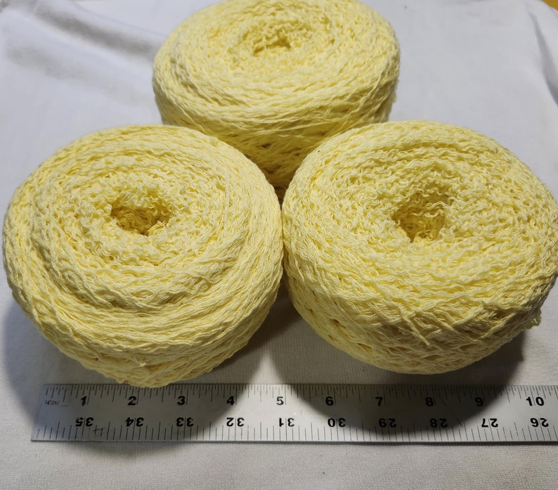 May include: Three round cakes of pale yellow yarn arranged in a triangular formation. A metal ruler is positioned at the base, showing that each yarn cake has a diameter of approximately 3.5 inches. The yarn has a textured, slightly crinkled appearance.
