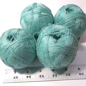 May include: Four balls of light teal yarn are arranged in a cluster above a metal ruler. The ruler shows the balls range from approximately 1.5 to 2 inches in diameter. The yarn has a textured, ribbon-like appearance with a matte finish.