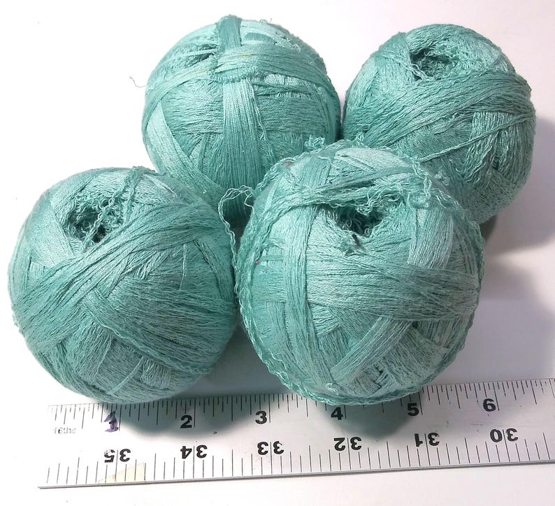 May include: Four balls of light teal yarn are arranged in a cluster above a metal ruler. The ruler shows the balls range from approximately 1.5 to 2 inches in diameter. The yarn has a textured, ribbon-like appearance with a matte finish.