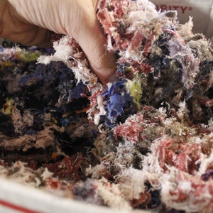 May include: A hand holds a mass of colorful fabric scraps inside a cardboard box. The scraps are a mix of red, blue, white, and yellow, creating a textured, multi-colored pile. The image suggests a craft or textile project.