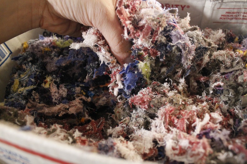 May include: A hand holds a mass of colorful fabric scraps inside a cardboard box. The scraps are a mix of red, blue, white, and yellow, creating a textured, multi-colored pile. The image suggests a craft or textile project.