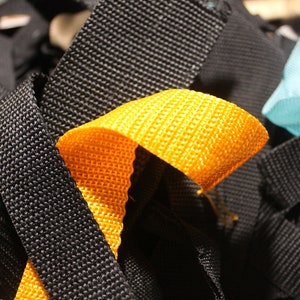 May include: A close-up of a pile of colorful woven straps. The straps are black, yellow, blue, and pink. The straps are woven with a textured pattern.