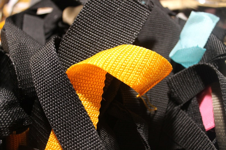 May include: A close-up of a pile of colorful woven straps. The straps are black, yellow, blue, and pink. The straps are woven with a textured pattern.