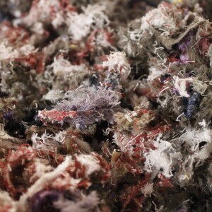 May include: Close-up of a pile of shredded fabric scraps in various colors. The scraps are primarily gray, with accents of red, white, purple, and yellow. The texture is rough and fibrous, suggesting a textile or craft material.
