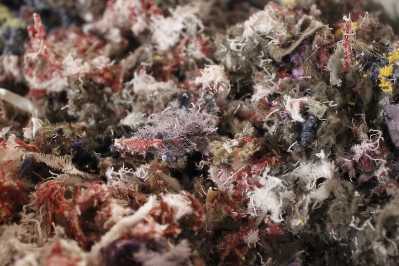 May include: Close-up of a pile of shredded fabric scraps in various colors. The scraps are primarily gray, with accents of red, white, purple, and yellow. The texture is rough and fibrous, suggesting a textile or craft material.
