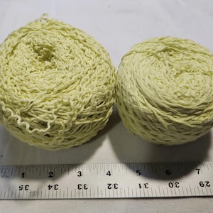 May include: Two balls of pale yellow yarn with a textured, crinkled appearance. A metal ruler is positioned below the yarn, showing the larger ball is approximately 3.5 inches wide and the smaller ball is approximately 2.5 inches wide.