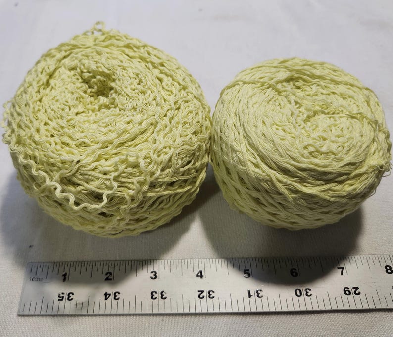 May include: Two balls of pale yellow yarn with a textured, crinkled appearance. A metal ruler is positioned below the yarn, showing the larger ball is approximately 3.5 inches wide and the smaller ball is approximately 2.5 inches wide.