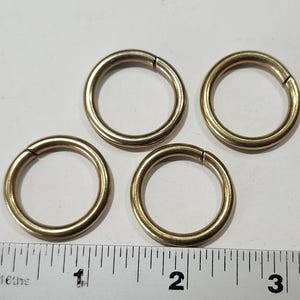 May include: Four gold-colored metal rings, each with a diameter of approximately 1 inch, are arranged on a white surface with a ruler for scale.