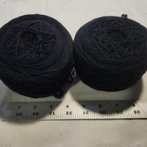 May include: Two round balls of black yarn positioned above a metal ruler. The ruler shows that each ball of yarn has a diameter of approximately 3.5 inches. The yarn is wound in a circular pattern, creating a textured surface on each ball.