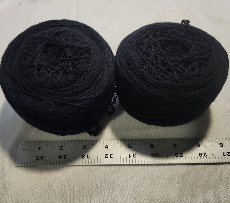 May include: Two round balls of black yarn positioned above a metal ruler. The ruler shows that each ball of yarn has a diameter of approximately 3.5 inches. The yarn is wound in a circular pattern, creating a textured surface on each ball.