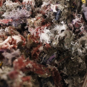 May include: Close-up of a mass of mixed textile scraps. The image shows a dense collection of shredded fabric in various colors, including red, purple, gray, and white. The texture appears soft and fibrous, suggesting a material used for stuffing or insulation.