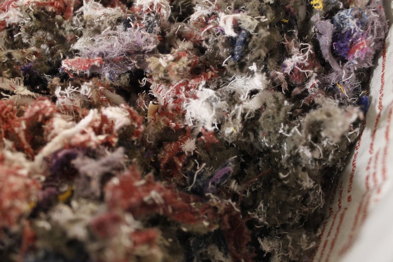 May include: Close-up of a mass of mixed textile scraps. The image shows a dense collection of shredded fabric in various colors, including red, purple, gray, and white. The texture appears soft and fibrous, suggesting a material used for stuffing or insulation.