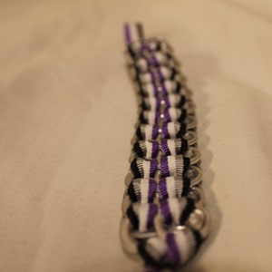 May include: A handmade bracelet crafted from recycled aluminum can tabs. The bracelet features a repeating pattern of silver tabs, black, white, and purple ribbons woven through the tabs. The bracelet is a unique accessory.