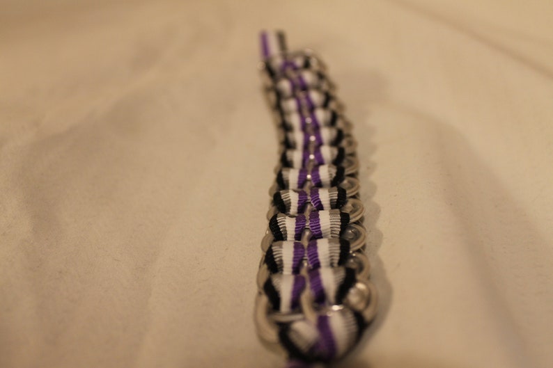 May include: A handmade bracelet crafted from recycled aluminum can tabs. The bracelet features a repeating pattern of silver tabs, black, white, and purple ribbons woven through the tabs. The bracelet is a unique accessory.