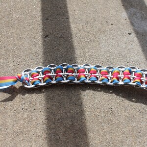 May include: A handmade bracelet featuring a chain of silver-colored metal loops, interwoven with colorful ribbons in shades of pink, yellow, and blue. The bracelet has a ribbon tie closure with the same color scheme, lying on a concrete surface.