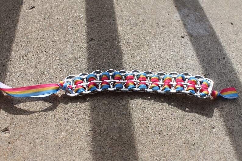 May include: A handmade bracelet featuring a chain of silver-colored metal loops, interwoven with colorful ribbons in shades of pink, yellow, and blue. The bracelet has a ribbon tie closure with the same color scheme, lying on a concrete surface.