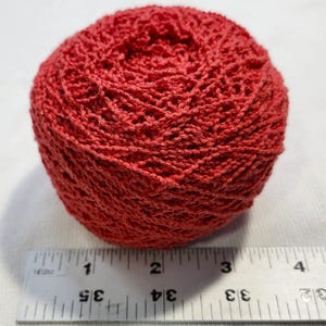 May include: A round ball of red yarn with a textured, knotted surface. A white ruler is positioned directly in front of the yarn, showing that the ball measures approximately 2.5 inches in diameter across its widest point.