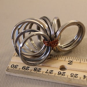 May include: A close-up of a set of silver key rings, some of which are interlocked. The key rings are arranged in a circular pattern and are resting on a wooden ruler marked with inches.