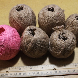 May include: Five balls of brown twine and one ball of pink twine are arranged in a group. A wooden ruler is positioned at the bottom of the frame, showing the balls measure approximately two inches in diameter across the scale.
