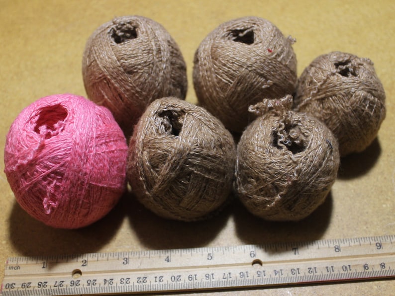 May include: Five balls of brown twine and one ball of pink twine are arranged in a group. A wooden ruler is positioned at the bottom of the frame, showing the balls measure approximately two inches in diameter across the scale.
