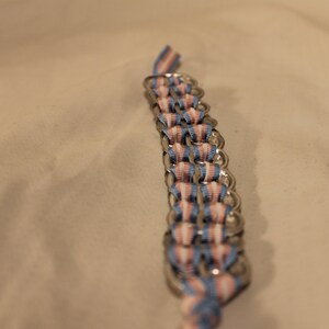 May include: A handmade bracelet composed of silver-colored metal rings and woven ribbons in blue, pink, and white. The bracelet is shown against a neutral background, highlighting the colors and craftsmanship of the piece.