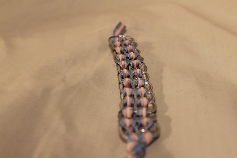 May include: A handmade bracelet composed of silver-colored metal rings and woven ribbons in blue, pink, and white. The bracelet is shown against a neutral background, highlighting the colors and craftsmanship of the piece.