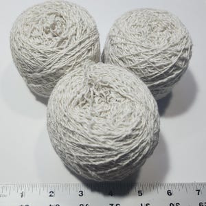 May include: Three round balls of off-white yarn arranged in a triangular formation. A ruler is positioned at the bottom, showing the width of the yarn balls spans from approximately 0.5 inches to 7.5 inches across the bottom edge of the image.