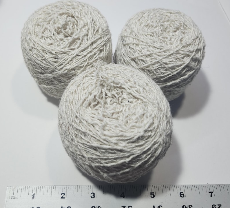 May include: Three round balls of off-white yarn arranged in a triangular formation. A ruler is positioned at the bottom, showing the width of the yarn balls spans from approximately 0.5 inches to 7.5 inches across the bottom edge of the image.