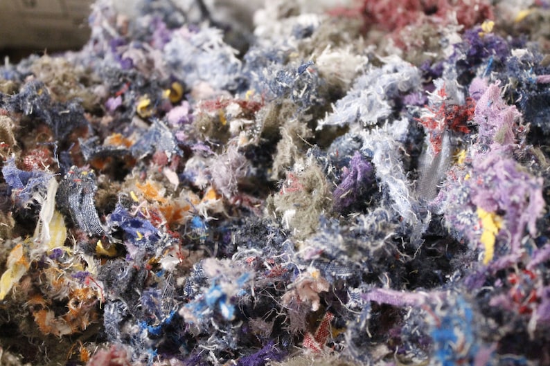 May include: Close-up of a pile of shredded fabric scraps in various colors. The scraps are a mix of blues, purples, reds, and yellows, with a textured, frayed appearance. The image suggests a textile recycling or craft project.