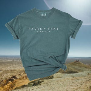 May include: A teal-colored t-shirt with the words "PAUSE + PRAY" and "JAMES 5:16" printed in white. The shirt is knotted at the bottom and has rolled-up sleeves. The background features a desert landscape under a blue sky.
