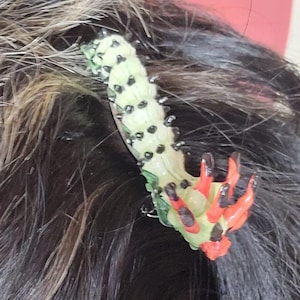 May include: A green and red plastic hair claw with black accents. The claw is shaped like a dragon's head with a long, thin body.