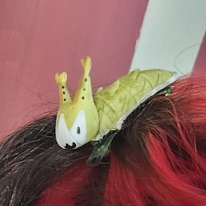 May include: A green and white caterpillar-shaped hair clip with black dots. The clip is in red and black hair.