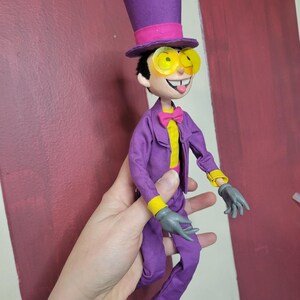 May include: A handmade doll dressed in a purple suit, yellow shirt, and pink bow tie. The doll has a purple top hat with a pink band, yellow-tinted glasses, and gray gloves. The doll's tongue is sticking out.