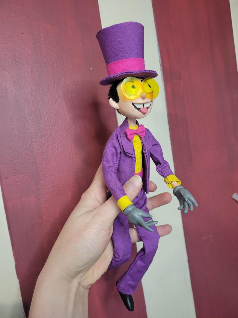 May include: A handmade doll dressed in a purple suit, yellow shirt, and pink bow tie. The doll has a purple top hat with a pink band, yellow-tinted glasses, and gray gloves. The doll's tongue is sticking out.