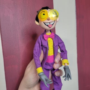 May include: A handmade doll dressed in a purple suit with yellow accents, a yellow shirt, and a pink bow tie. The doll has yellow-rimmed glasses and gray gloves. The doll's face has a wide smile and its tongue is sticking out.