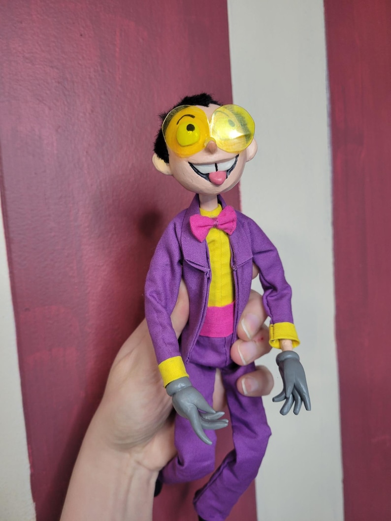 May include: A handmade doll dressed in a purple suit with yellow accents, a yellow shirt, and a pink bow tie. The doll has yellow-rimmed glasses and gray gloves. The doll's face has a wide smile and its tongue is sticking out.