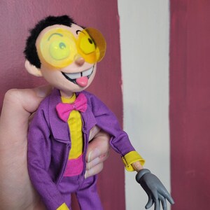 May include: A handmade doll with a cartoonish design. The doll wears a purple suit with yellow accents, a pink bow tie, and large yellow-tinted glasses. The doll has a smiling face with its tongue sticking out and gray gloves.