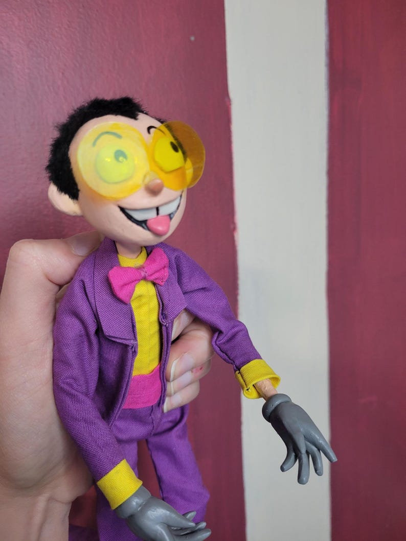 May include: A handmade doll with a cartoonish design. The doll wears a purple suit with yellow accents, a pink bow tie, and large yellow-tinted glasses. The doll has a smiling face with its tongue sticking out and gray gloves.